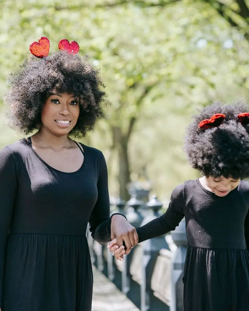 A mother and her young daughter holding hands and smiling on a tree-lined park path